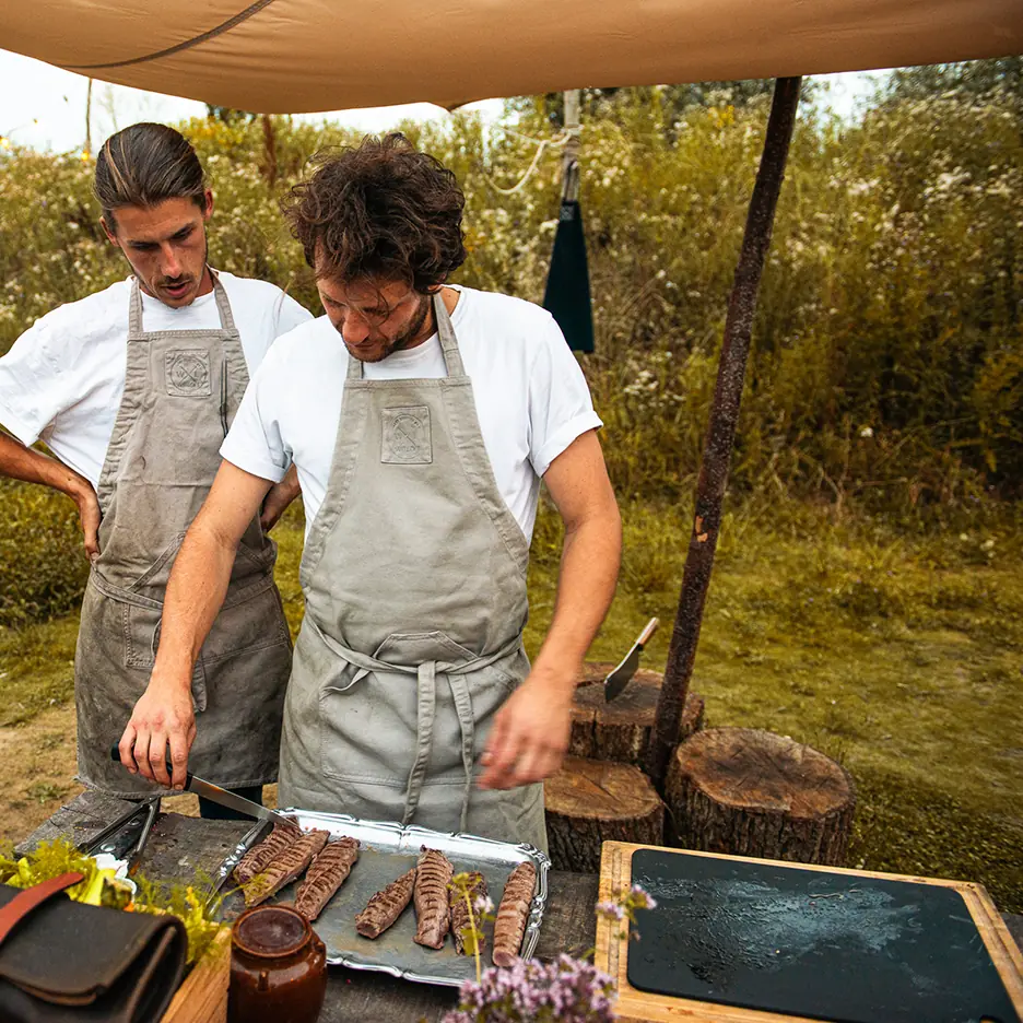 Twee koks in grijze schorten bereiden vlees op een tray in een buitenkeuken.