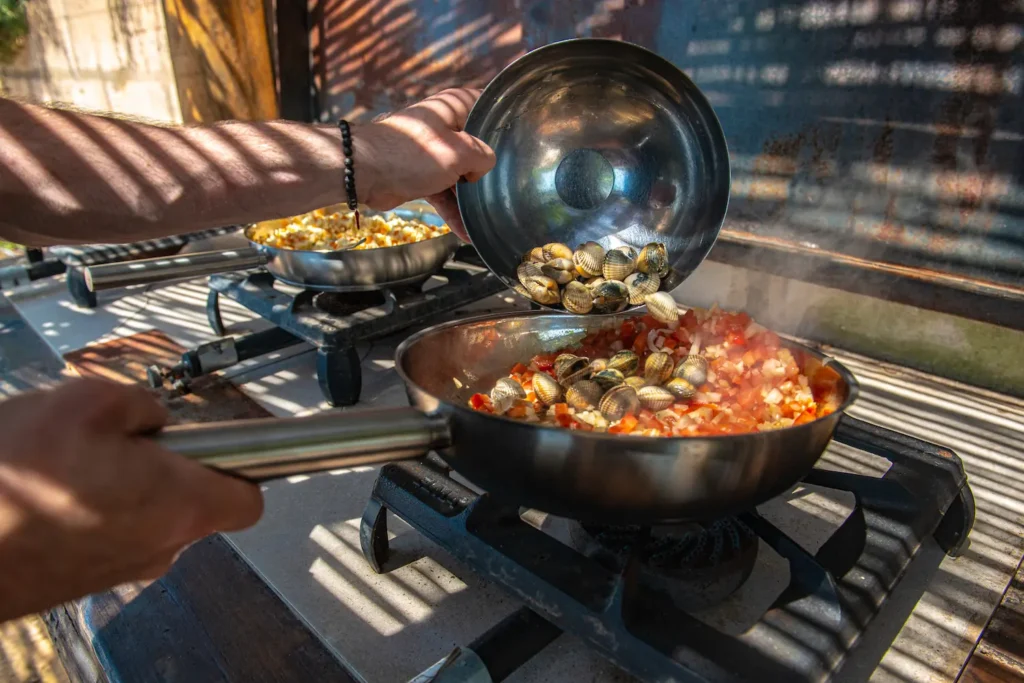 Een pan met venusschelpen wordt toegevoegd aan een mengsel van tomaten en uien op een buitenkooktoestel.